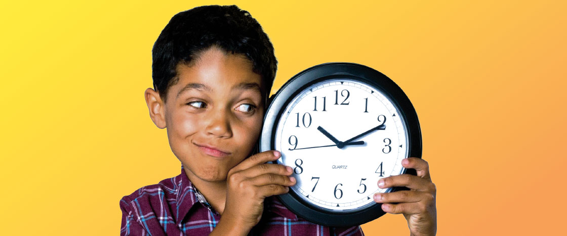 A boy wearing a plaid shirt looking at the round clock he is holding up next to his face